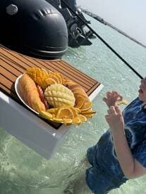 A beautifully arranged fruit platter sits on the edge of a boat while a child enjoys a snack in the shallow water — perfect for refreshing treats during a day on the water.