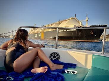 A woman enjoys a scenic view from a small boat, gazing at a large LNG ship in the background — a perfect moment of relaxation on the water.