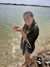 A smiling woman holds a small shell while standing in shallow water, showcasing a delightful moment of human-animal interaction during a beach outing.