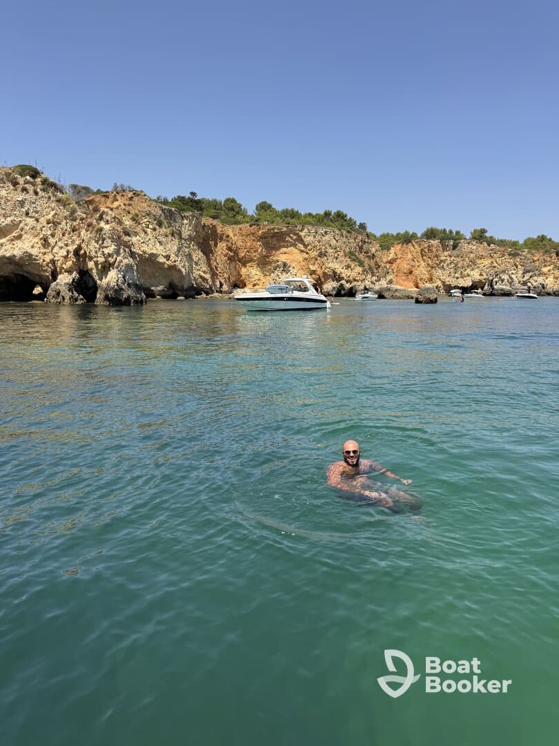 A man enjoys a refreshing swim in clear waters near a picturesque coastline, with a boat anchored nearby — a perfect scene for summer adventures.