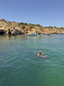A man enjoys a refreshing swim in clear waters near a picturesque coastline, with a boat anchored nearby — a perfect scene for summer adventures.