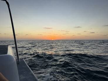 A serene sunset view from the side of a boat, showcasing the tranquil ocean waves and the vibrant colors of the sky as the sun sets on the horizon.