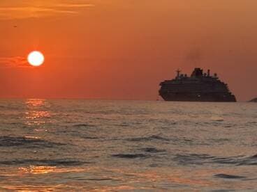 A stunning sunset casts a warm glow over a cruise ship silhouetted against the horizon, creating a picturesque maritime scene.