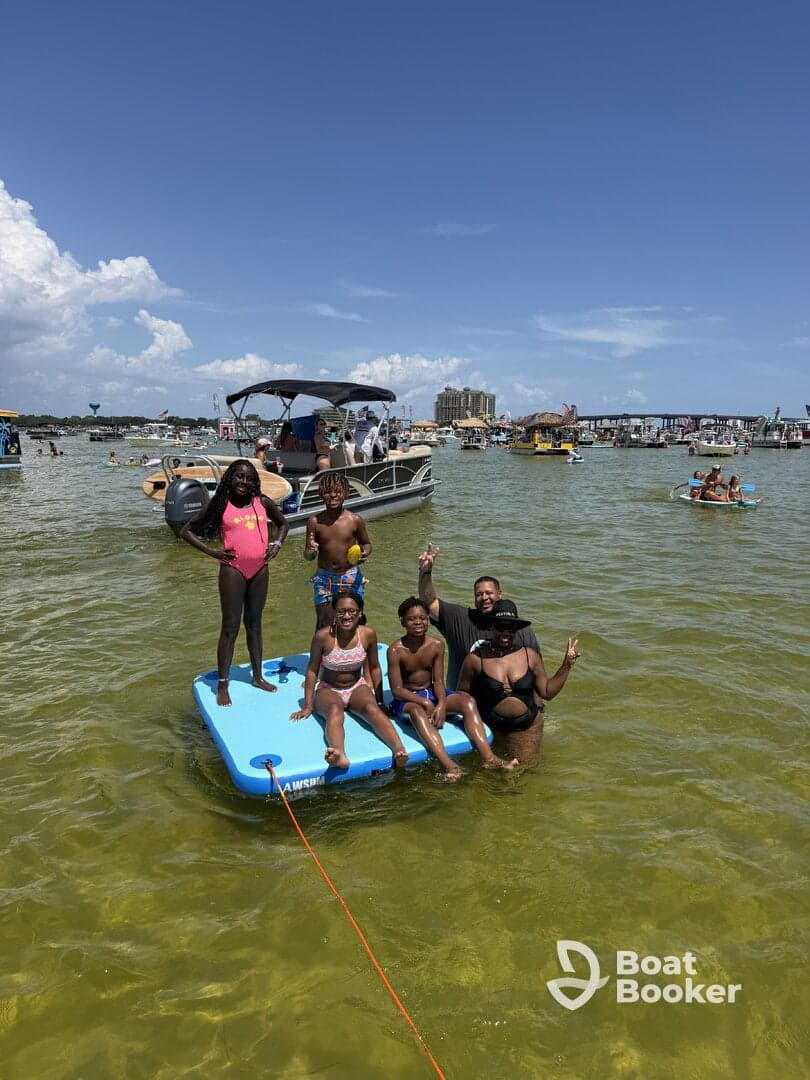 A joyful group of family members enjoys a sunny day on the water, relaxing on a floating mat while a pontoon boat is anchored nearby — perfect for summer fun and family bonding.
