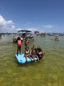 A joyful group of family members enjoys a sunny day on the water, relaxing on a floating mat while a pontoon boat is anchored nearby — perfect for summer fun and family bonding.