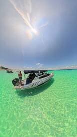 A boat anchored in crystal-clear turquoise waters with a person standing on deck, surrounded by a vibrant beach scene — perfect for summer adventures and water activities.