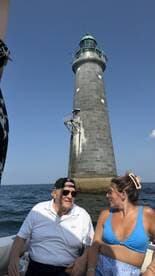 A couple enjoys a scenic boat ride near a historic lighthouse, capturing a moment of connection against a beautiful ocean backdrop.