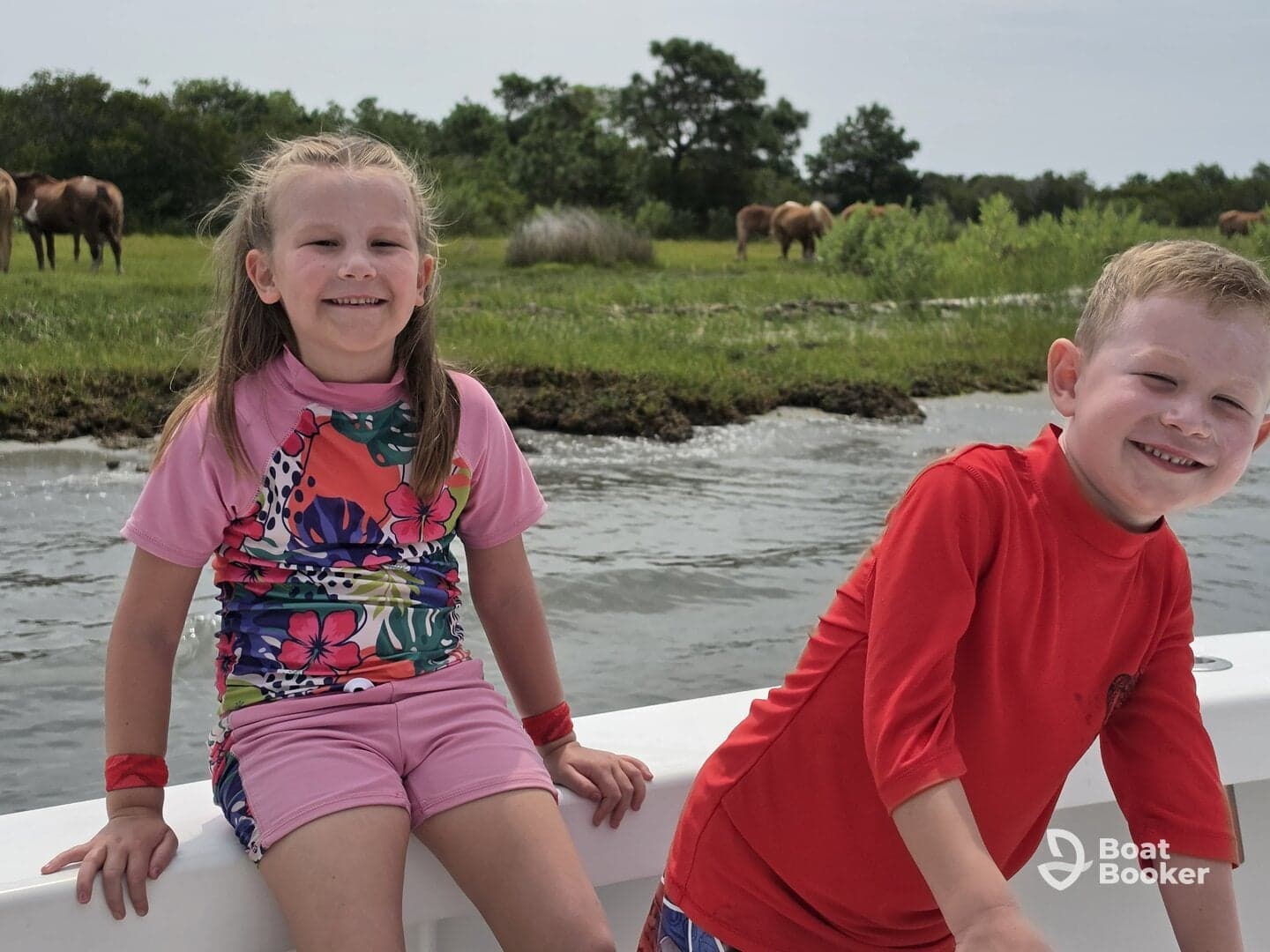 Two children enjoy a fun day on the boat, smiling as they take in the scenic view of wild horses grazing nearby — a perfect family adventure on the water.