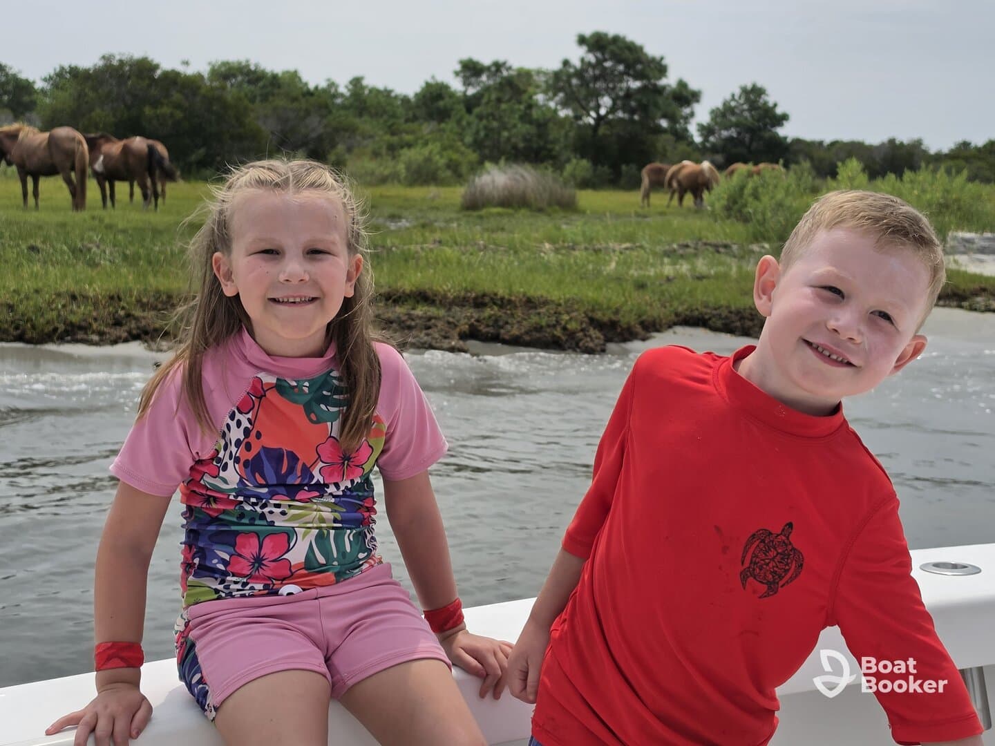 Two children smile from the boat as wild horses graze in the background, capturing a joyful family moment on the water.