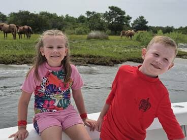 Two children smile from the boat as wild horses graze in the background, capturing a joyful family moment on the water.