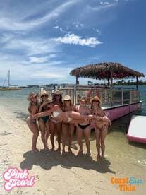 A lively group of friends celebrates on the beach with a fun pose, showcasing their vibrant energy and the beautiful backdrop of the Pink Pearl tiki boat.
