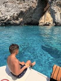 A young boy enjoys a sunny day by the water, dangling his feet in the clear turquoise sea while surrounded by fish — a perfect moment for family fun and snorkeling adventures.