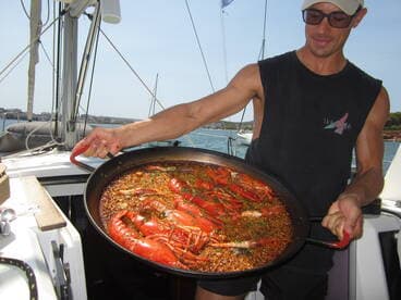 A chef presents a delicious seafood paella on a sailboat, showcasing onboard catering options for a memorable dining experience at sea.