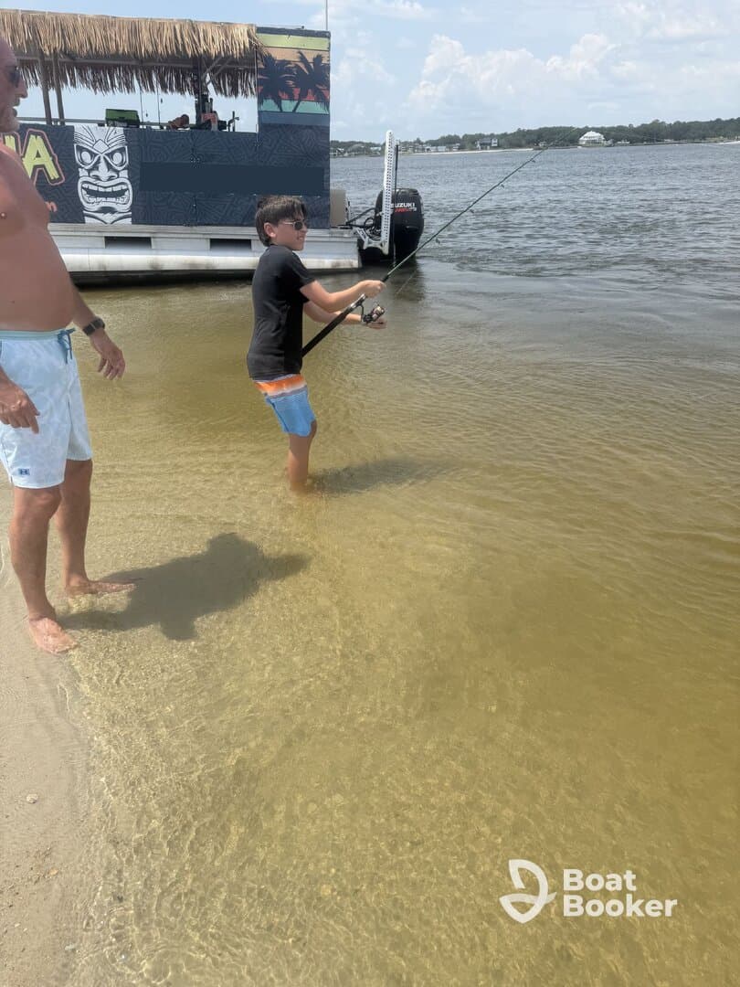 A child enjoys fishing in shallow water while an adult watches nearby, creating a memorable family moment by the pontoon boat.