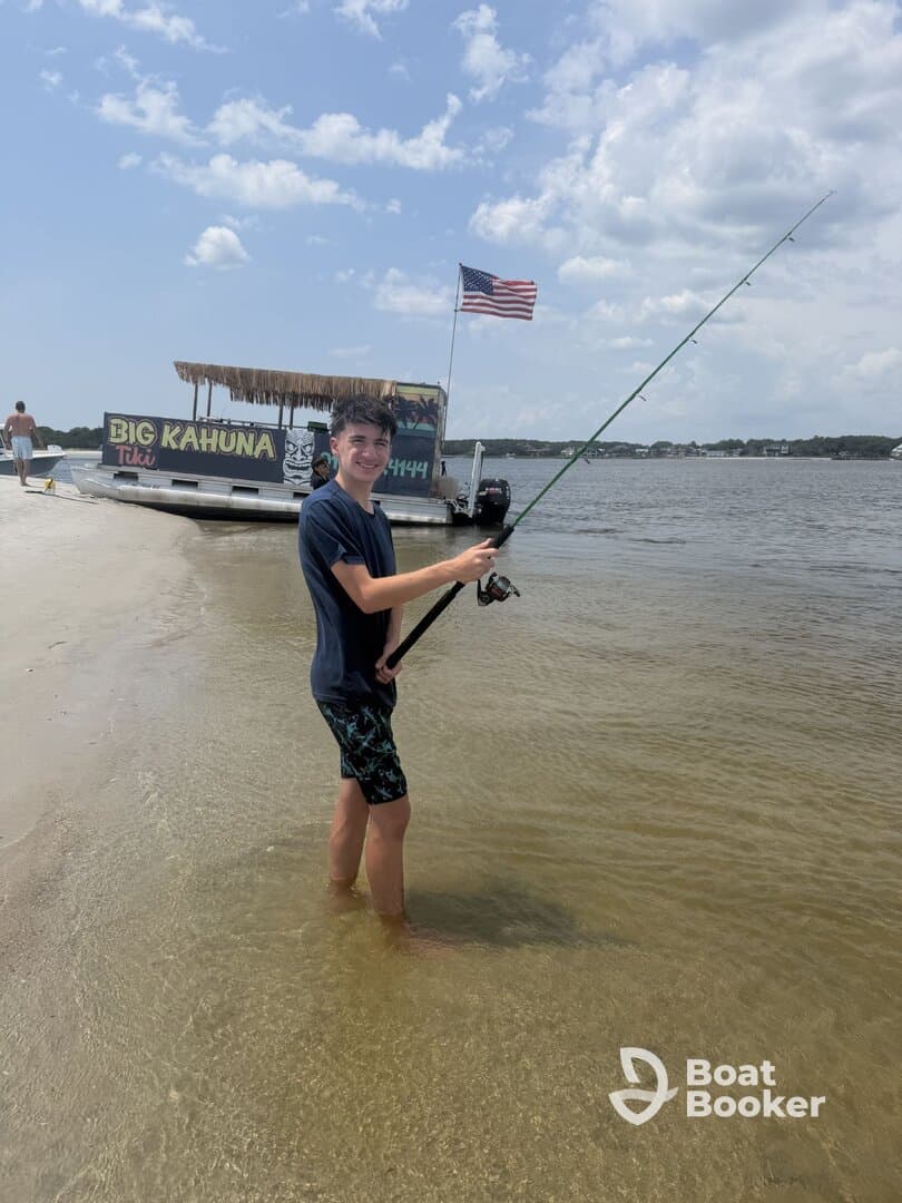 A young boy enjoys fishing in shallow waters near the 'Big Kahuna' tiki boat, capturing the essence of summer fun and outdoor adventure.