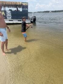 A child enjoys fishing in shallow water while an adult watches nearby, creating a memorable family moment by the pontoon boat.