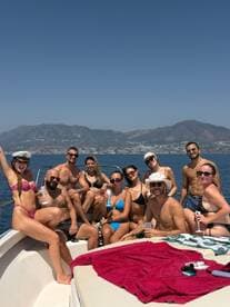 A lively group enjoys a fun day on the water, celebrating with drinks and sunbathing aboard a boat, surrounded by stunning coastal views.