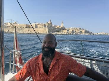A smiling man enjoys a scenic boat ride with a stunning cityscape in the background, perfect for capturing memorable vacation moments.
