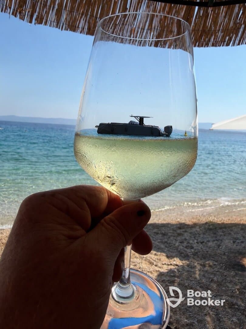 A hand holding a glass of wine with a boat reflected in it, set against a beach backdrop. The unique perspective creates an artistic view of a relaxing day by the water.