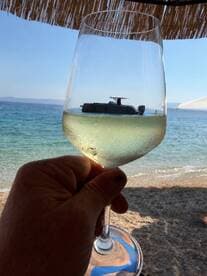A hand holding a glass of wine with a boat reflected in it, set against a beach backdrop. The unique perspective creates an artistic view of a relaxing day by the water.