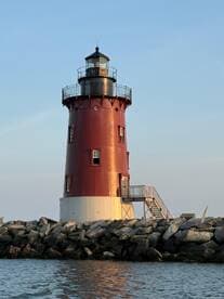 A striking red lighthouse stands tall against a clear sky, marking the entrance to a harbor. Its unique design and coastal setting make it a picturesque landmark.