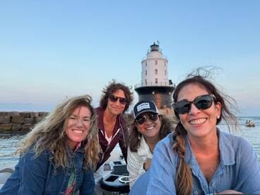 A cheerful group of friends poses for a selfie on a boat with a lighthouse in the background, capturing a memorable moment on the water.