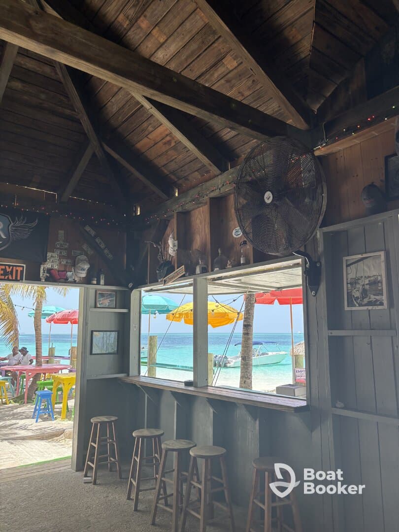 Interior view of a beachside bar with wooden seating and a view of colorful umbrellas and boats outside, perfect for relaxing after a day on the water.