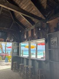 Interior view of a beachside bar with wooden seating and a view of colorful umbrellas and boats outside, perfect for relaxing after a day on the water.