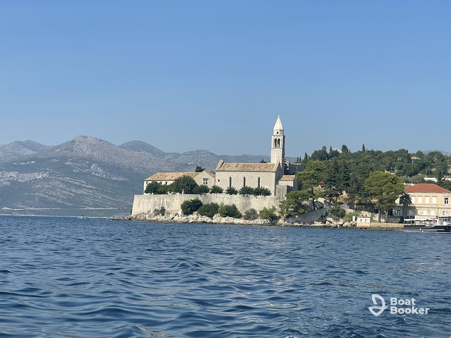 A picturesque view of a coastal landmark with a historic church, framed by majestic mountains and clear blue waters — a perfect representation of the stunning scenery found while boating.