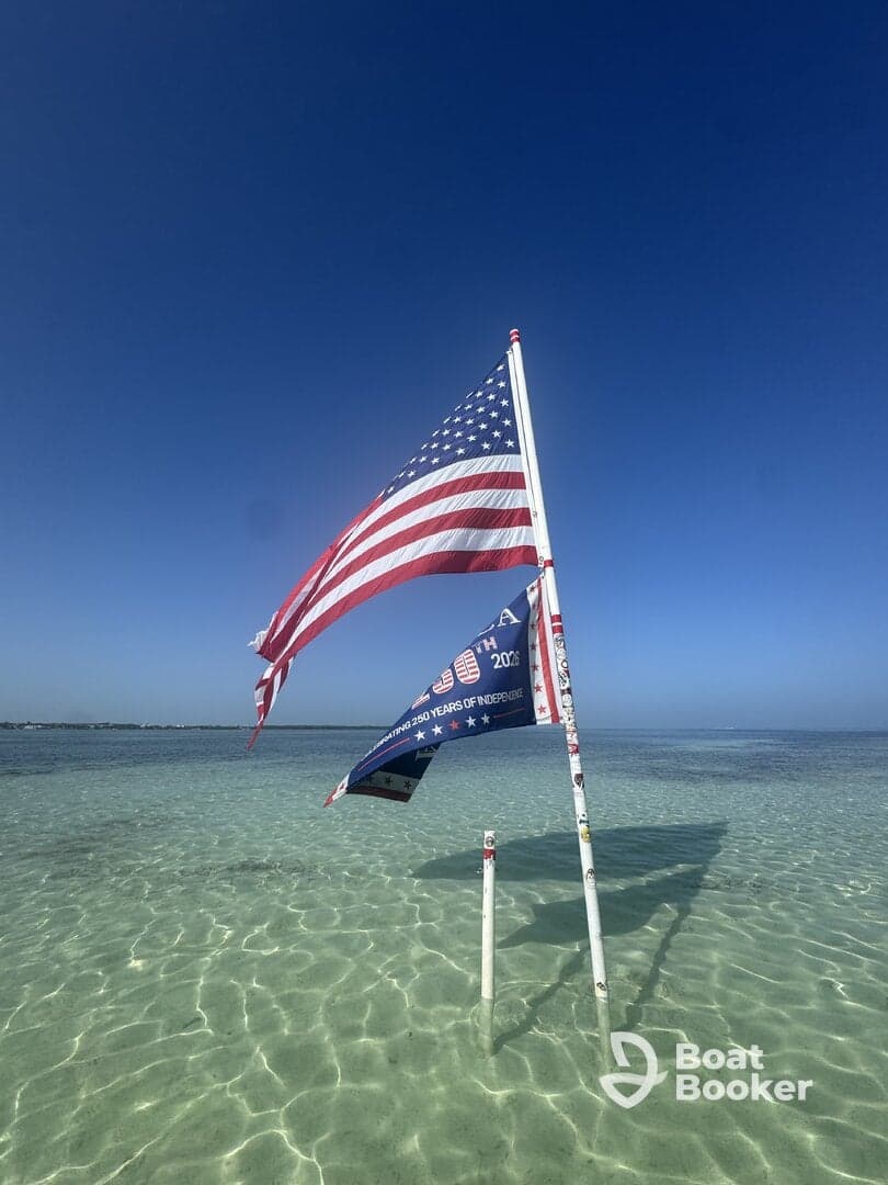 A flagpole displaying an American flag and another flag in shallow water, with a clear blue sky above. The scene captures a serene coastal atmosphere.