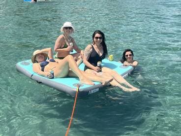 A fun group of friends relaxes on a floating mat in crystal-clear waters, enjoying drinks and sunshine — the perfect setting for a summer getaway.