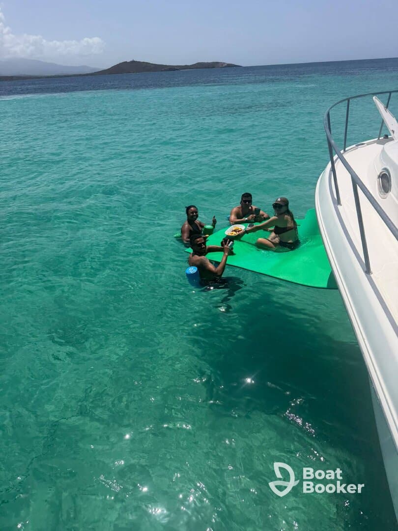 A lively group enjoys food and drinks on a floating mat in crystal-clear waters, perfect for a fun day out on the boat.