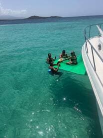 A lively group enjoys food and drinks on a floating mat in crystal-clear waters, perfect for a fun day out on the boat.