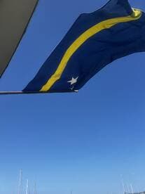 A flag waving against a clear blue sky, with sailboats visible in the background — a serene maritime scene.