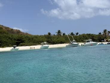 A scenic view of several boats anchored near a beach, with a group of people enjoying the sun and sand — perfect for a relaxing day on the water.