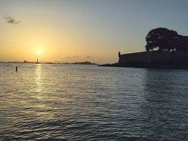 A stunning sunset view over the harbor, featuring a silhouette of a historic fort and a sailboat in the distance — perfect for capturing the beauty of coastal destinations.