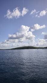 A scenic view of a calm water body with clouds overhead and a distant island, creating a tranquil atmosphere.