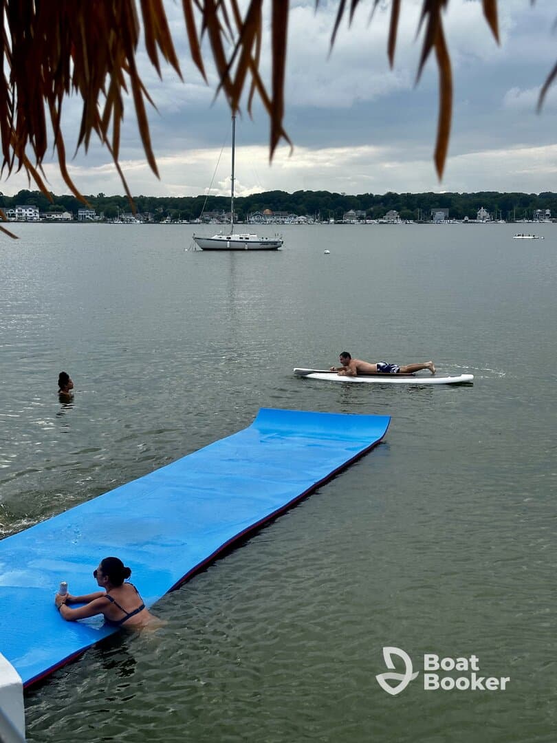 A lively scene featuring friends enjoying water activities, including paddleboarding and relaxing on a floating mat, with a sailboat anchored in the background.