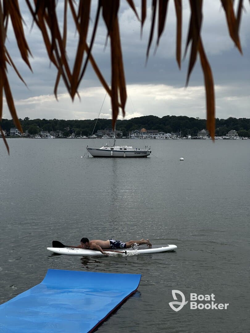 A man paddleboards in calm waters with a sailboat anchored in the background, showcasing a serene day on the water.
