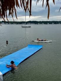 A lively scene featuring friends enjoying water activities, including paddleboarding and relaxing on a floating mat, with a sailboat anchored in the background.