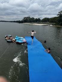 A lively scene of friends enjoying a day on the water, with one jumping off a floating platform while others relax on inflatable loungers — perfect for summer fun and water activities.
