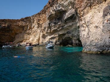 A swimmer enjoys the clear waters near a stunning coastal cave, with boats anchored nearby, creating a perfect scene for a summer adventure.
