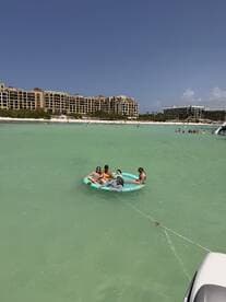 A lively group enjoys a fun day on a floating mat in the clear waters, surrounded by a beautiful beach backdrop — perfect for summer adventures.