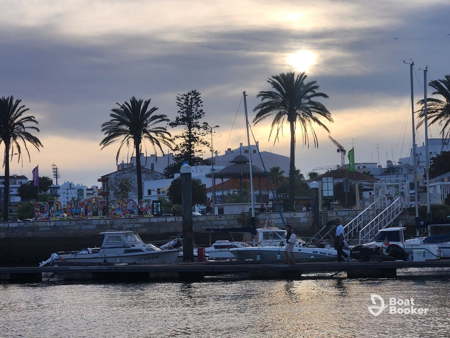 A picturesque sunset view at the harbor, featuring boats and palm trees, with people enjoying the waterfront atmosphere.