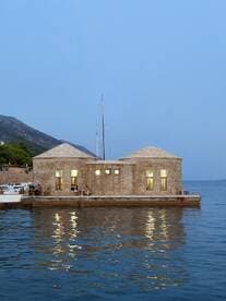 A picturesque scene featuring a stone building by the water, with a sailboat nearby and people visible, creating a serene atmosphere at dusk.