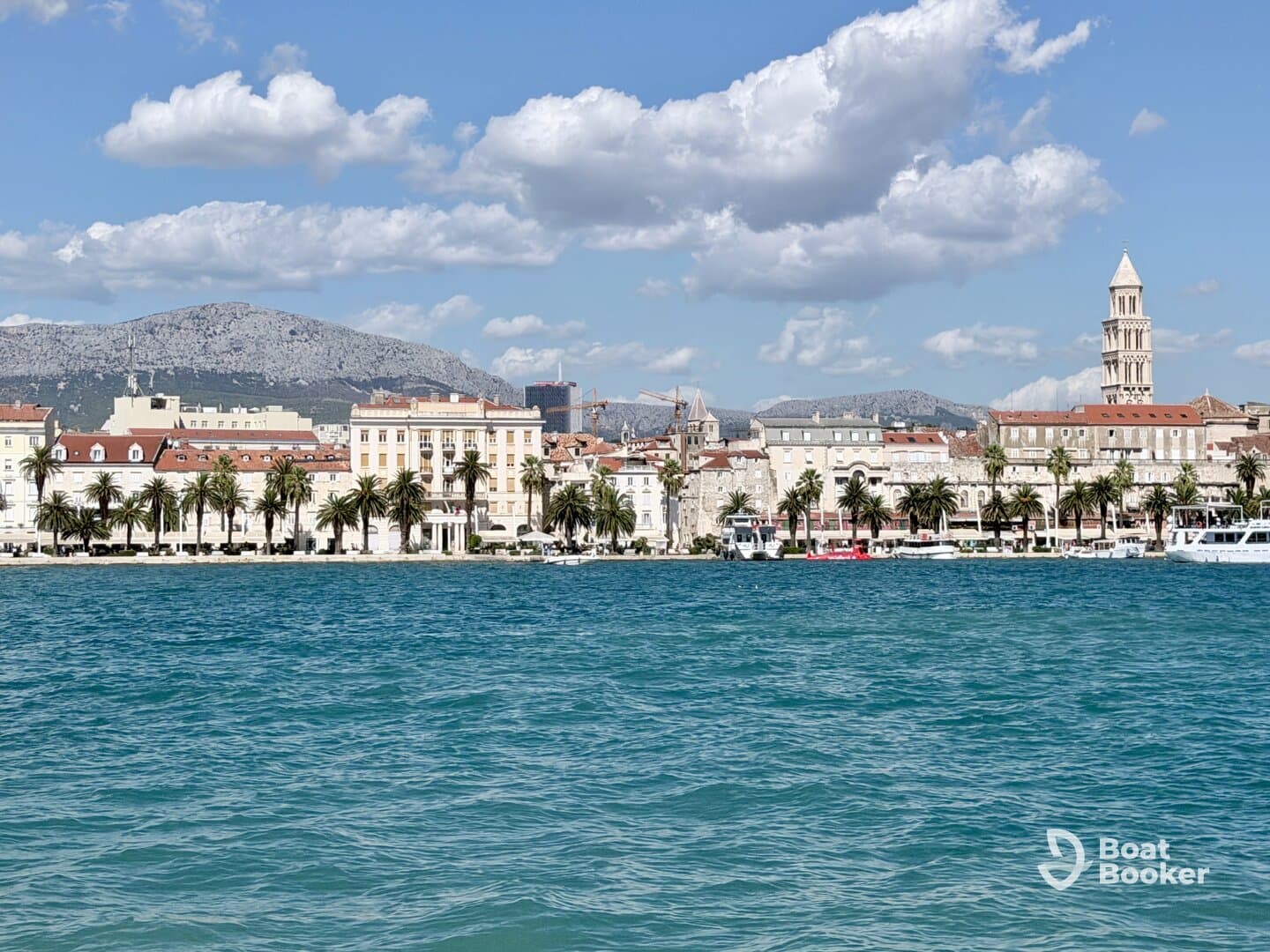 A picturesque view of the coastline featuring palm trees and historic architecture, set against a backdrop of mountains and a clear blue sky — a perfect representation of a vibrant harbor scene.