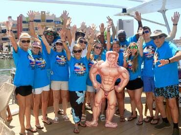 A lively group of friends celebrates aboard a boat, showcasing their fun spirit with matching shirts and a humorous inflatable figure — perfect for memorable party experiences on the water.