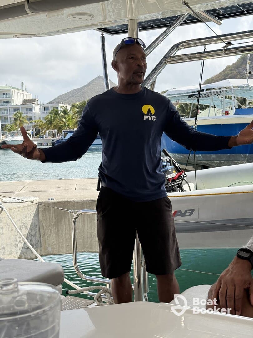 A man gestures while speaking on a boat, likely providing information or instructions, set against a scenic marina backdrop.