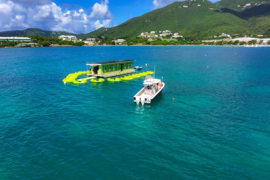 There's a small boat anchored by a floating bar with yellow seating, so you’ll get unique boat dining on clear blue water. Green hills and buildings are in the background.