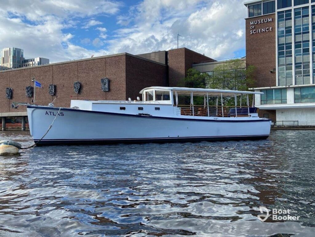 A white boat named ATLAS is docked on the water near the Museum of Science building in Boston, with city buildings visible in the background.